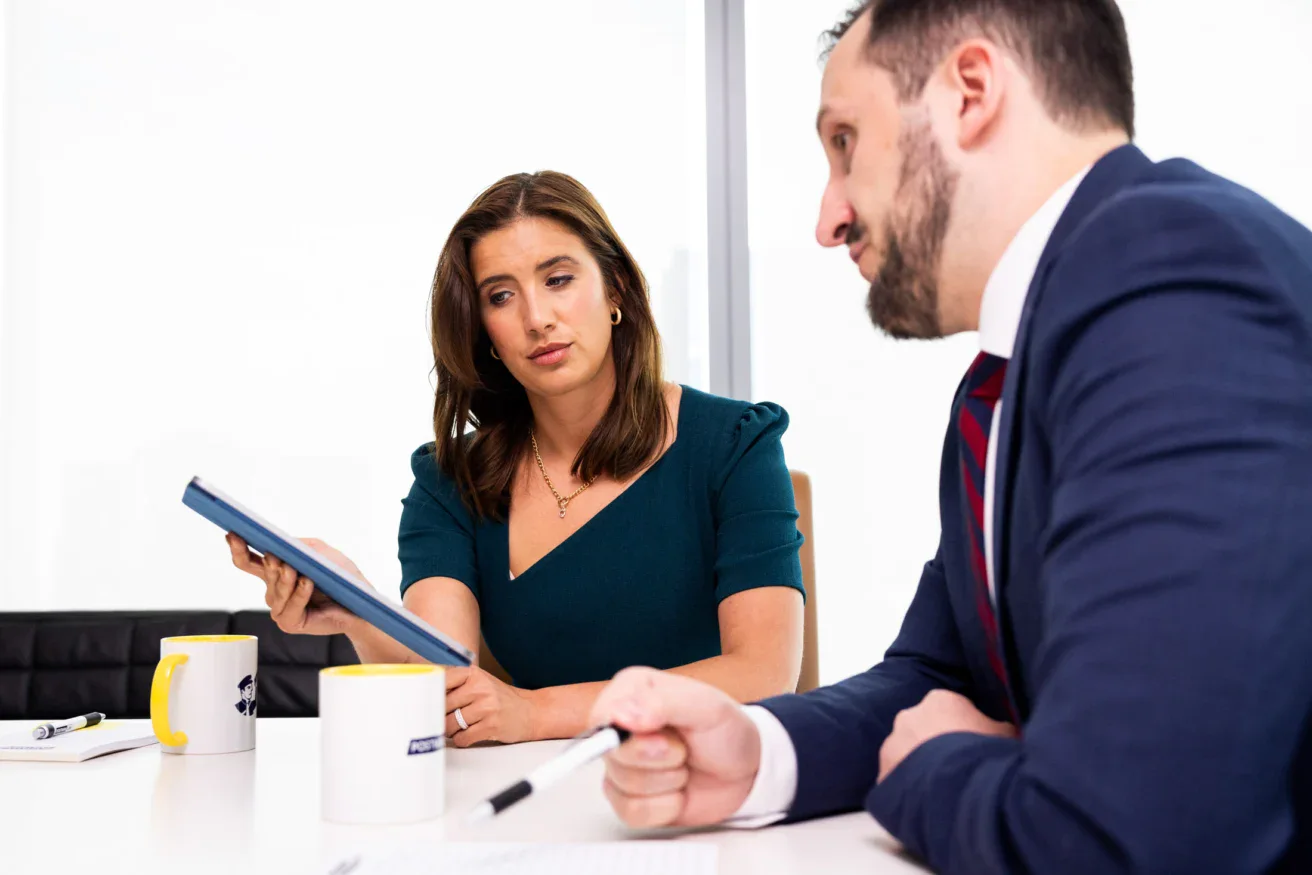 Two people in business attire sit at a table with notebooks, mugs, and papers; the woman holds a tablet and looks at the man, who is writing.