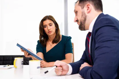 Two people in business attire sit at a table with notebooks, mugs, and papers; the woman holds a tablet and looks at the man, who is writing.
