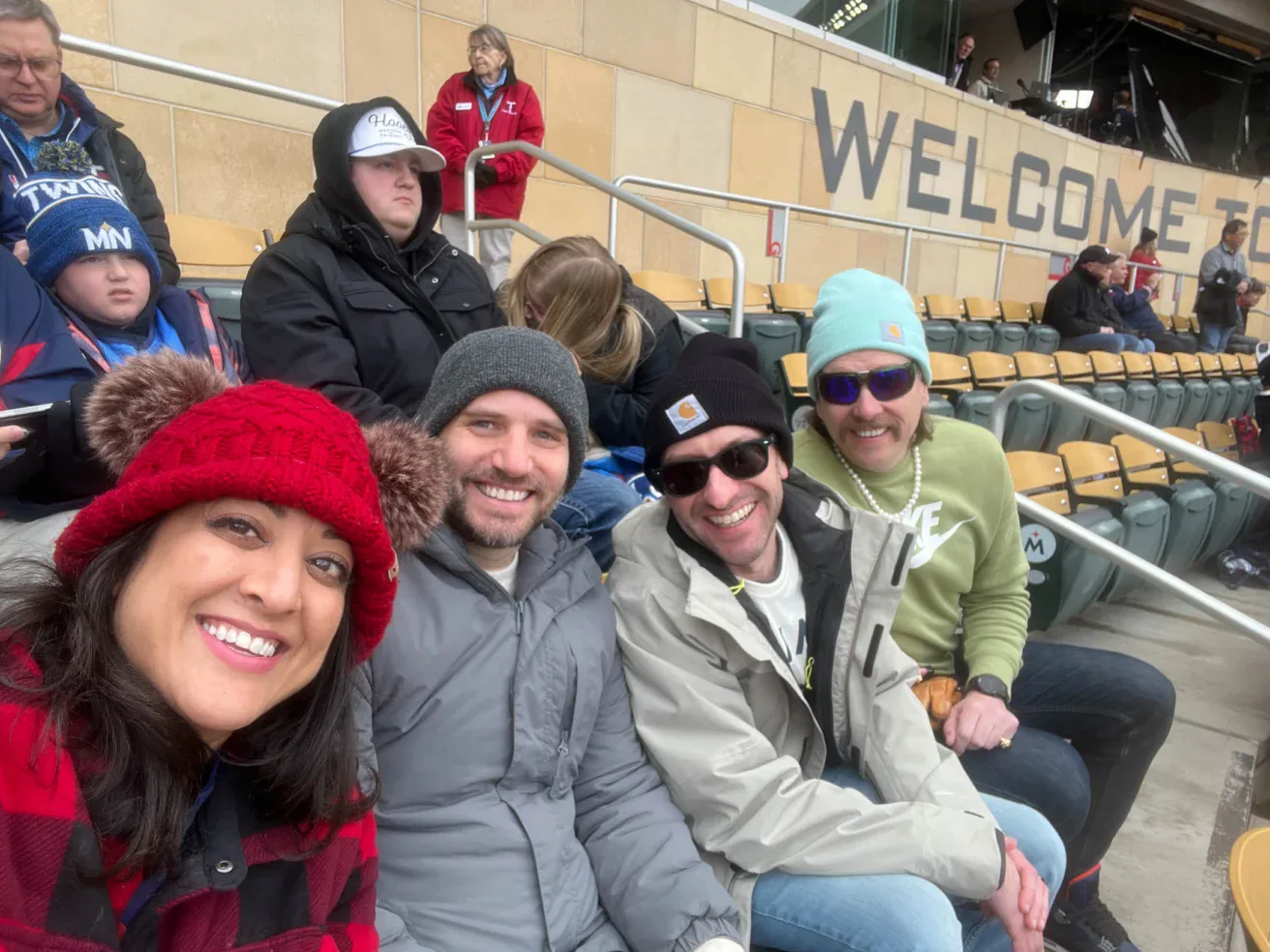 Four adults sit in stadium seats, dressed in winter clothing, smiling at the camera. Other attendees and seats are visible in the background near a “WELCOME TO” sign.