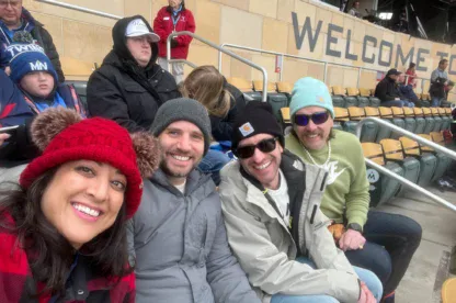 Four adults sit in stadium seats, dressed in winter clothing, smiling at the camera. Other attendees and seats are visible in the background near a “WELCOME TO” sign.