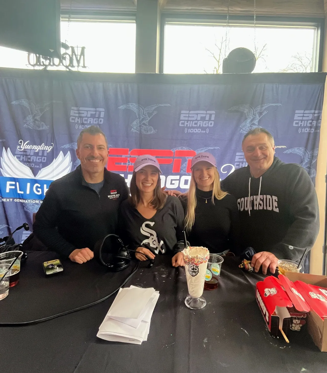 Four people stand and smile at a table with food and drinks in front of an ESPN Chicago banner. Two women wear matching light purple hats; one man wears a Southside shirt.