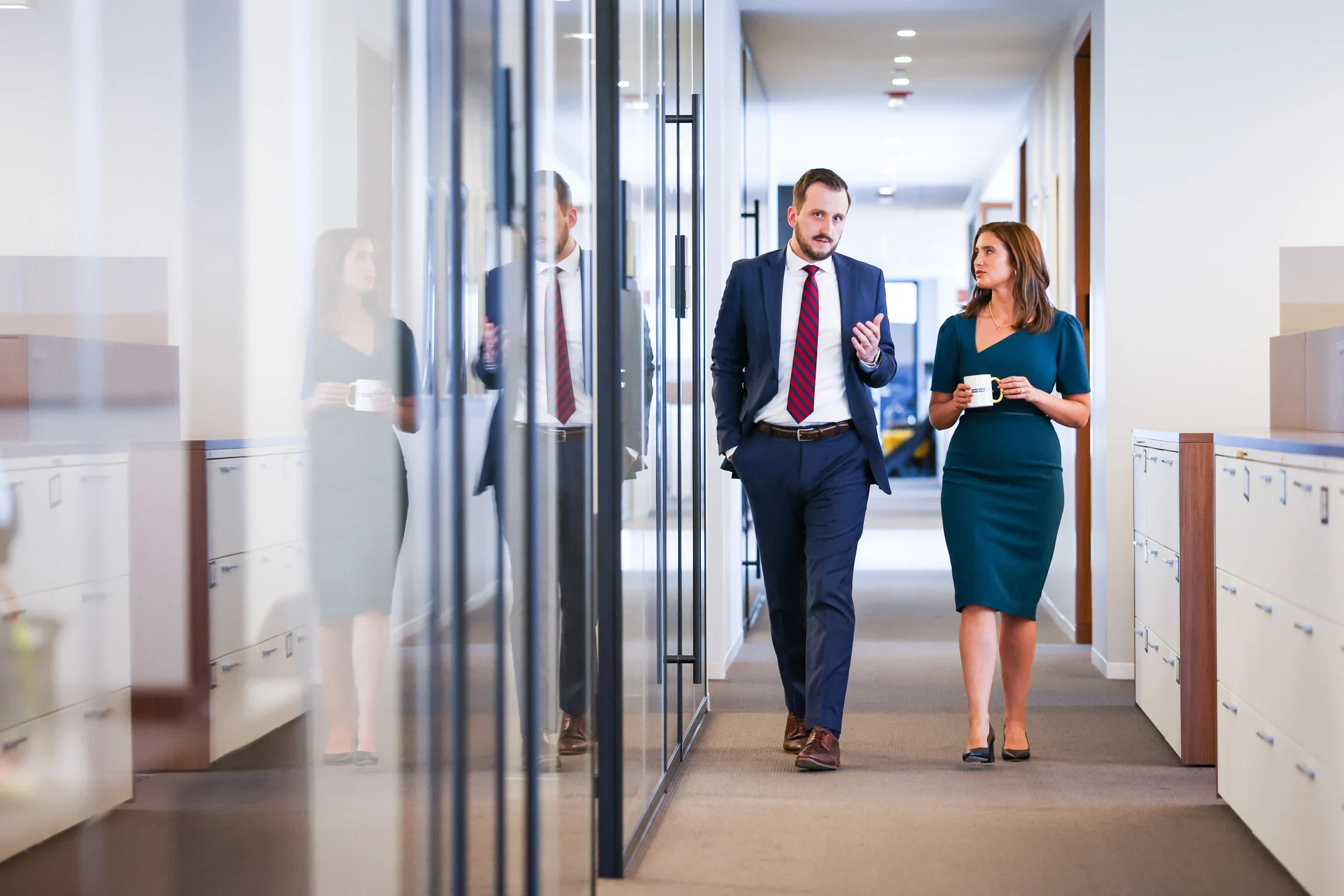 Two professionally dressed people walk and talk in an office hallway, holding coffee cups and appearing engaged in conversation.
