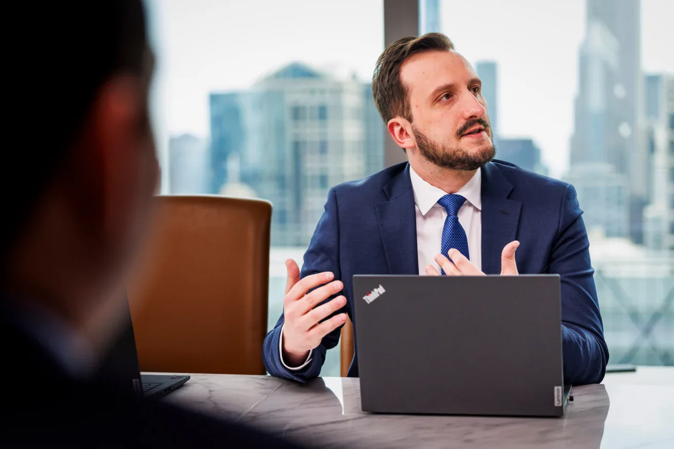A man in a suit sits at a table with a laptop, gesturing as he speaks during a business meeting in an office with city views.