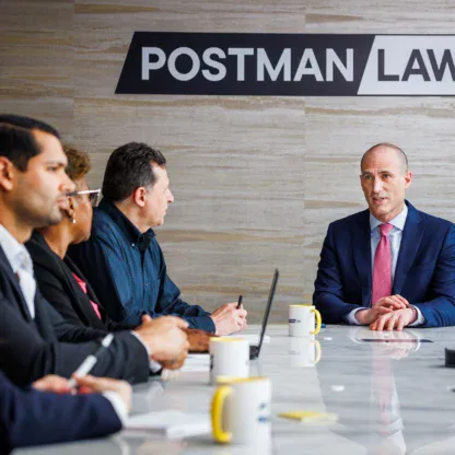 A group of professionals in business attire meet at a conference table under a sign that reads "POSTMAN LAW.