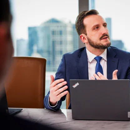 A man in a suit sits at a table with a laptop, gesturing as he speaks during a business meeting in an office with city views.