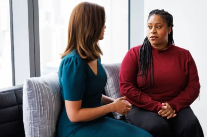 Two women sit on a couch in conversation; one wears a teal dress and the other a red long-sleeve top. They appear engaged in a serious discussion.