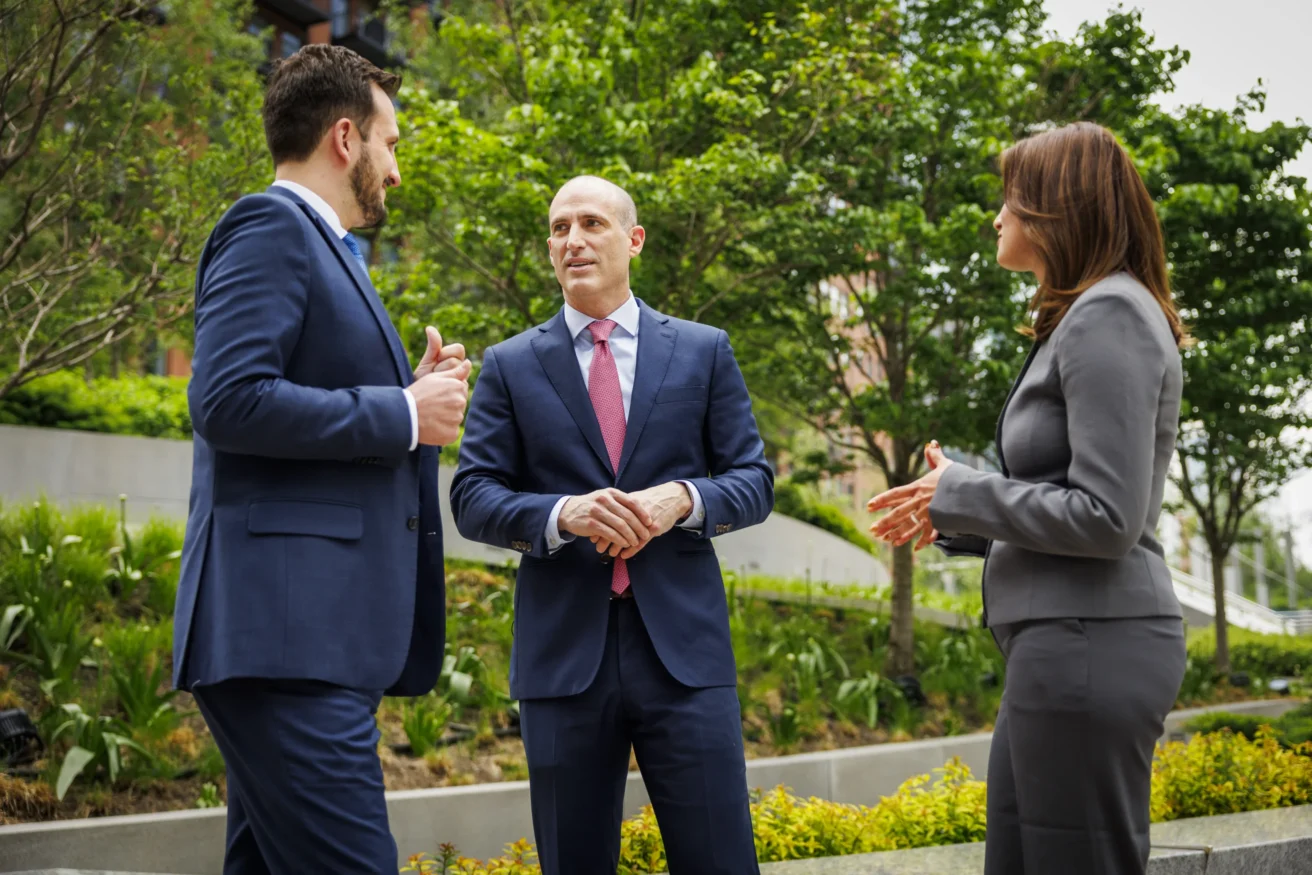 Three people in business attire stand outdoors having a discussion, with greenery and a building visible in the background.
