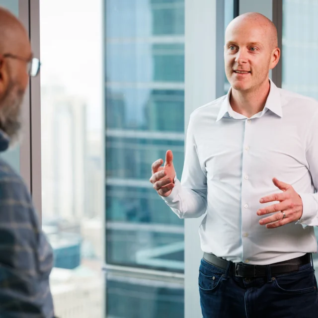 Two men in conversation by office windows in a high-rise building, with one man gesturing while the other listens.