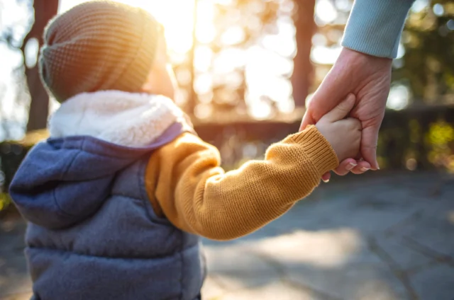 A small child in a warm jacket and hat holds an adult's hand while walking outdoors on a sunny day.