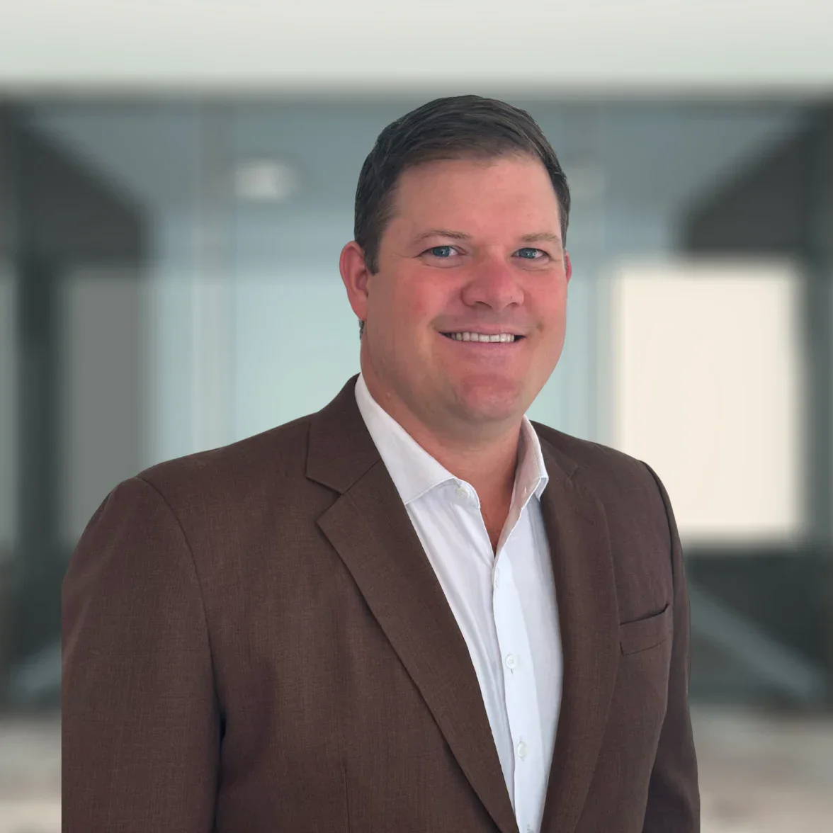 Un hombre con traje de chaqueta marrón y camisa blanca sonríe ante un fondo de oficina difuminado.