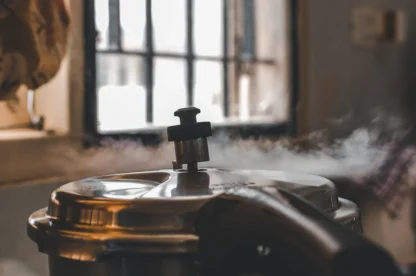 Steam escapes from the valve of a stainless steel pressure cooker in a kitchen, with a barred window and some utensils visible in the background.