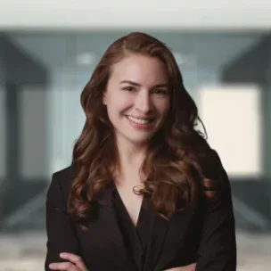 A woman with long brown hair, wearing a black blazer, stands smiling with her arms crossed in a modern office setting.