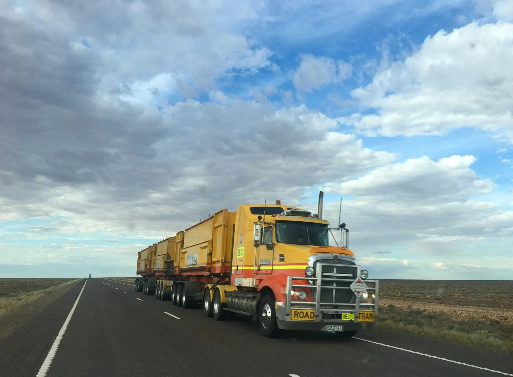 A large yellow road train truck with multiple trailers drives along an empty highway under a partly cloudy sky in a flat, open landscape.