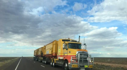 A large yellow road train truck with multiple trailers drives along an empty highway under a partly cloudy sky in a flat, open landscape.