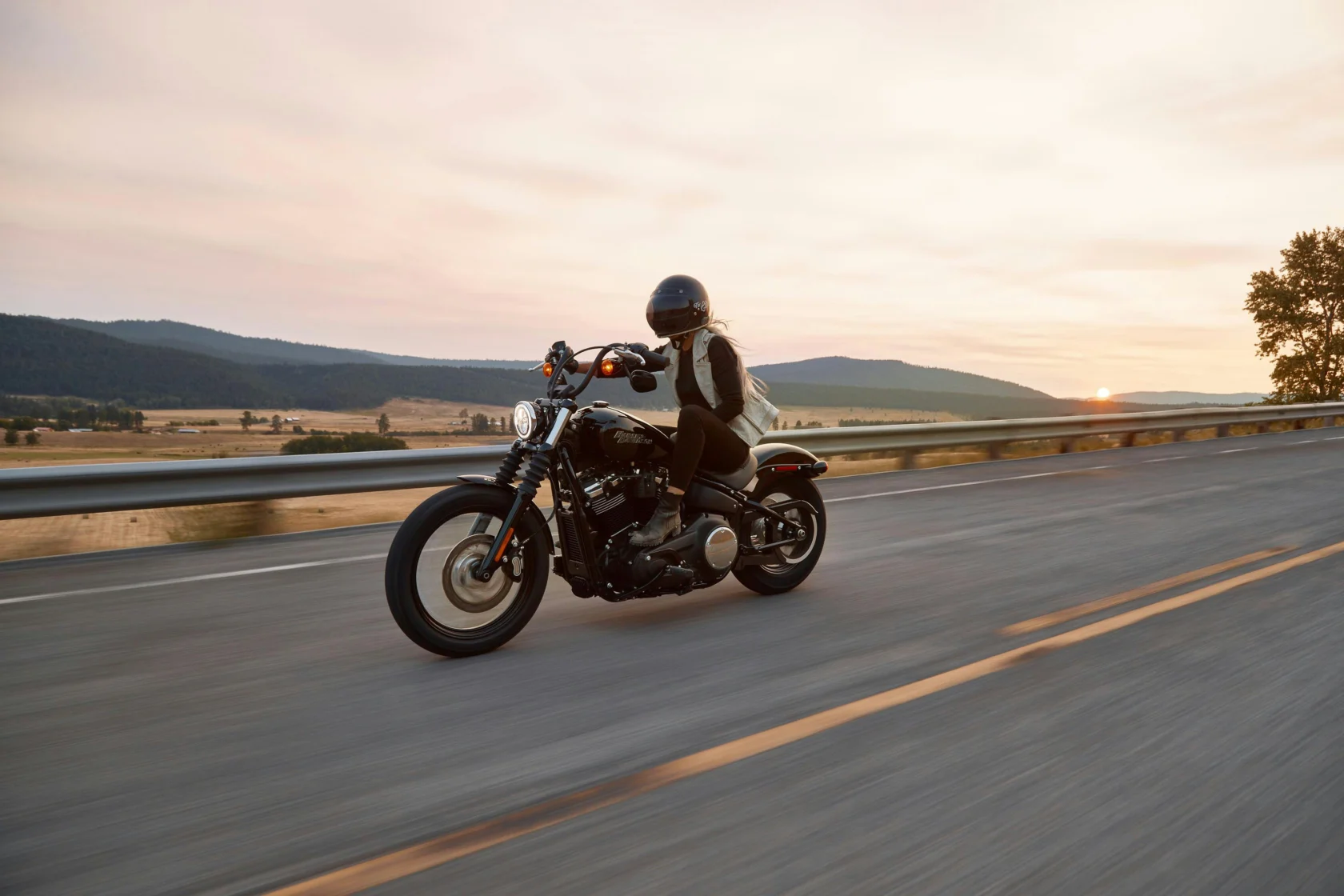A person in a helmet rides a black motorcycle on an open highway at sunset with hills and fields in the background.