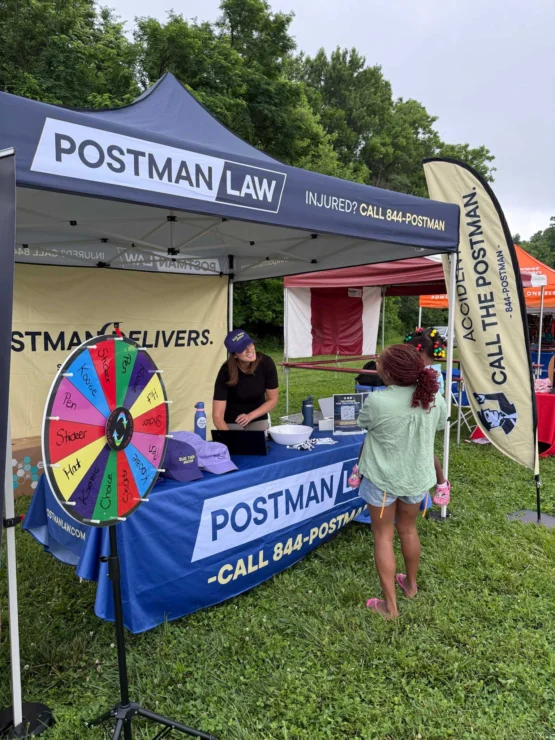 A woman sits at a Postman Law booth with a prize wheel, talking to a young girl at an outdoor event. The booth has banners and promotional items on display.